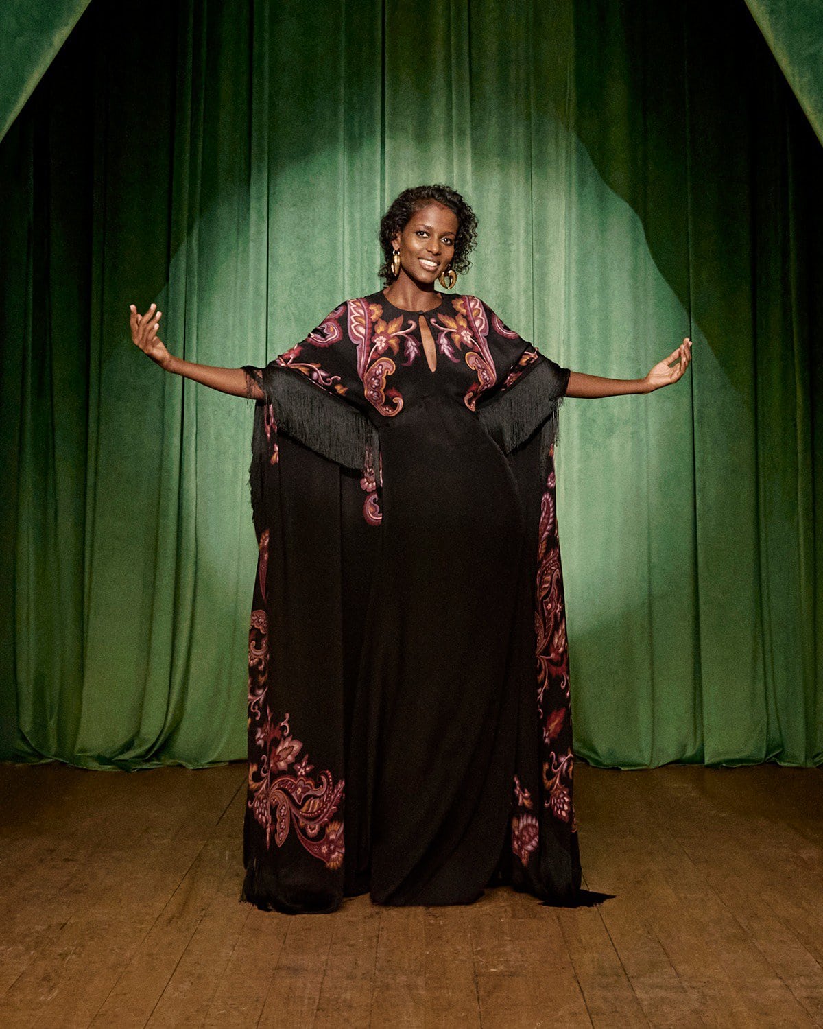 Model wearing a black patterned gown with fringe details standing on a stage with green velvet curtains.