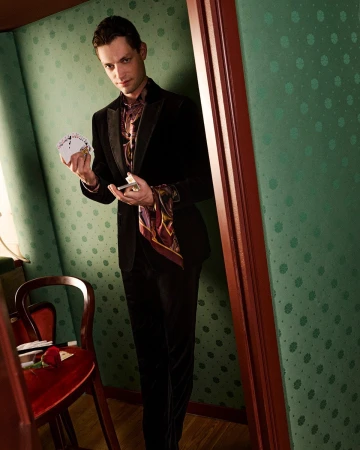 Man in dark velvet suit holding playing cards standing in a green patterned room