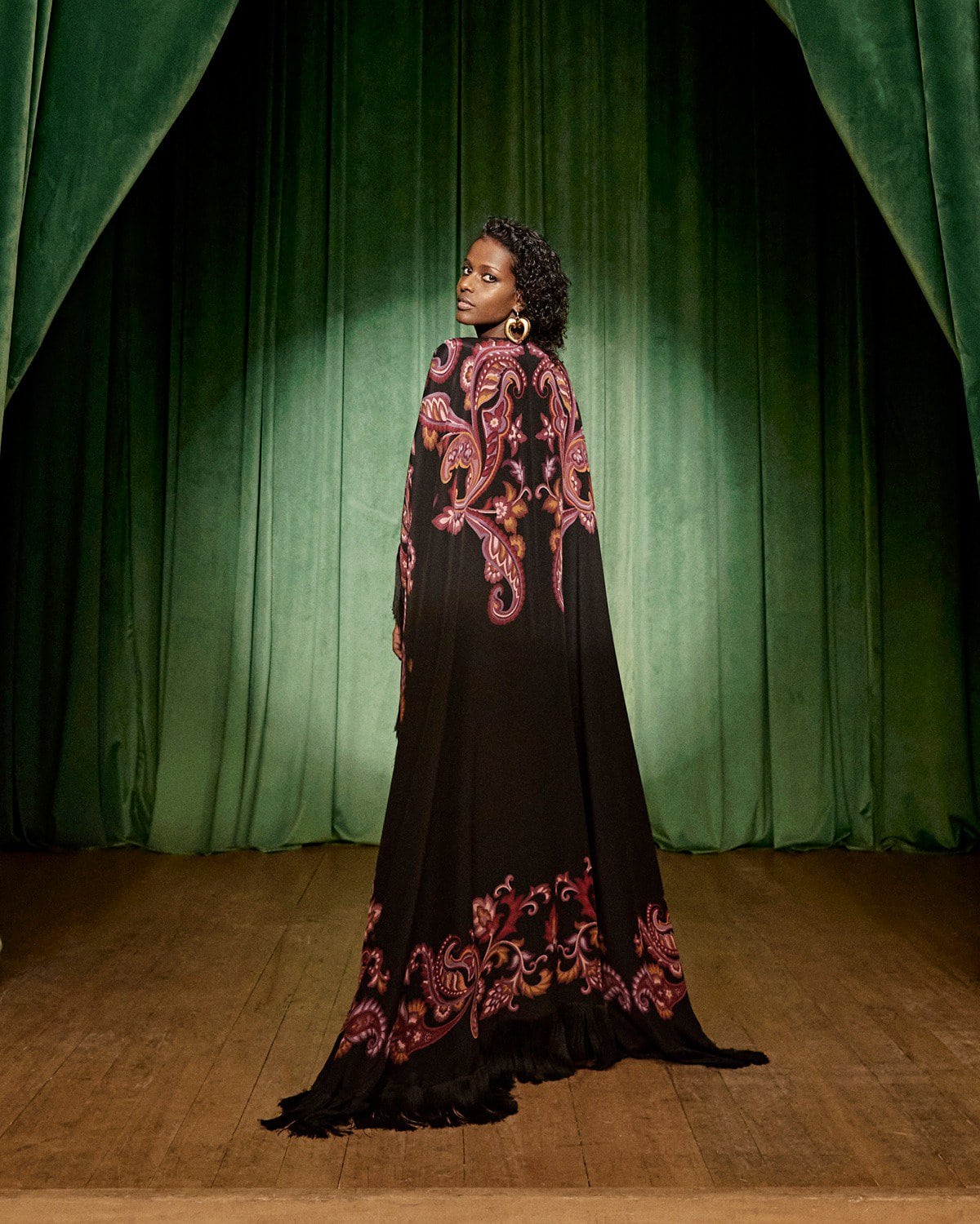 Model wearing a long black patterned cape standing on a stage with green curtains under a spotlight