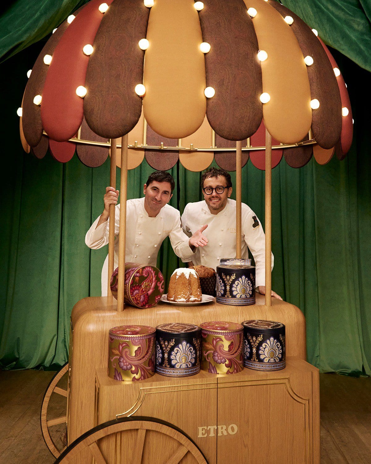Two men in white chef coats standing behind a wooden cart with a festive umbrella and panettone cakes for Etro Holiday campaign.