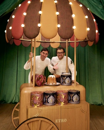 Two men in white chef coats standing behind a wooden cart with a festive umbrella and panettone cakes for Etro Holiday campaign.