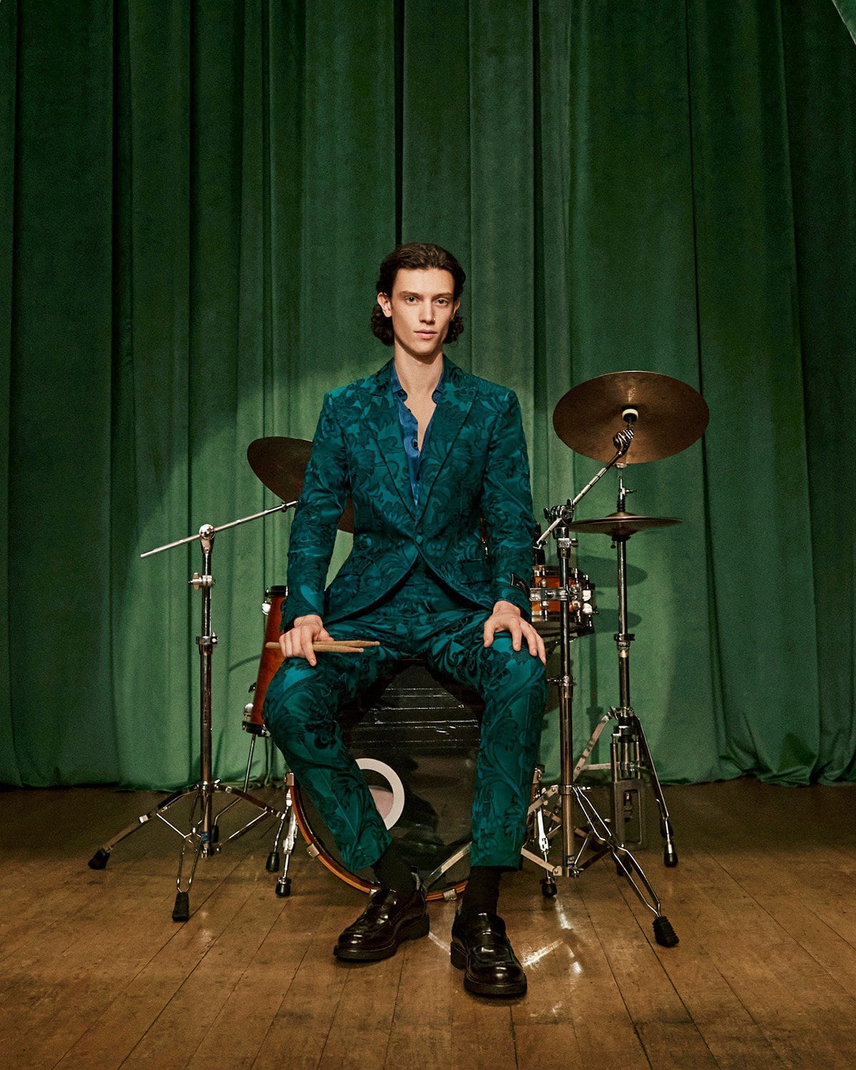 A man in a green patterned suit seated behind a drum kit against a green draped theatrical curtain.