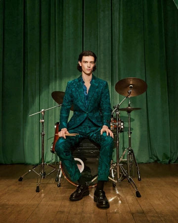 A man in a green patterned suit seated behind a drum kit against a green draped theatrical curtain.