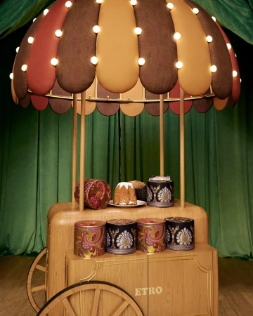 Wooden dessert cart with a striped canopy and lit bulbs displaying ornate packaging boxes and a panettone against a green curtain.