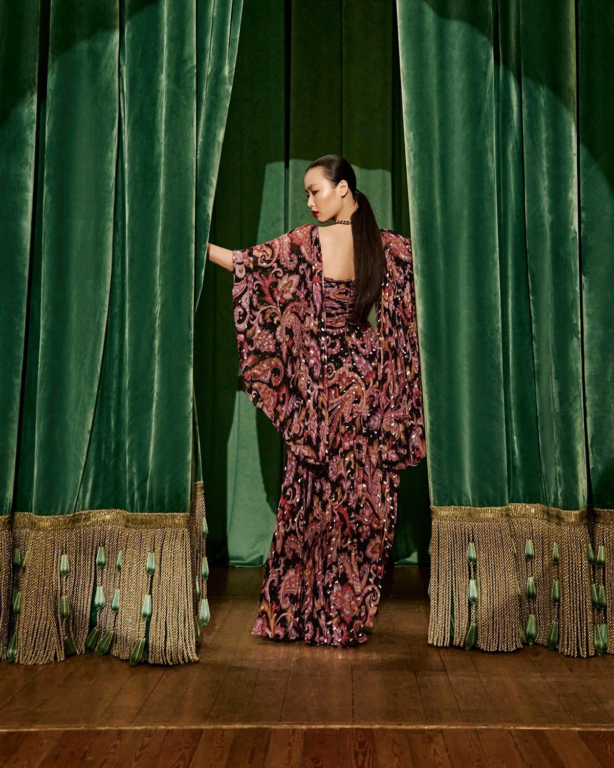 Model standing on a wooden stage parting heavy green velvet stage curtains in an Etro campaign.