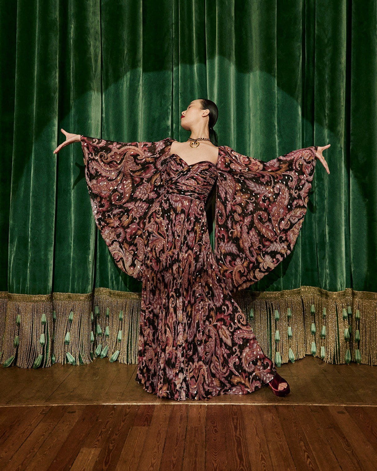 Model in patterned gown with dramatic draped sleeves standing on a wooden stage before heavy green velvet theater curtains.