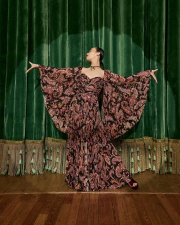 Model in patterned gown with dramatic draped sleeves standing on a wooden stage before heavy green velvet theater curtains.