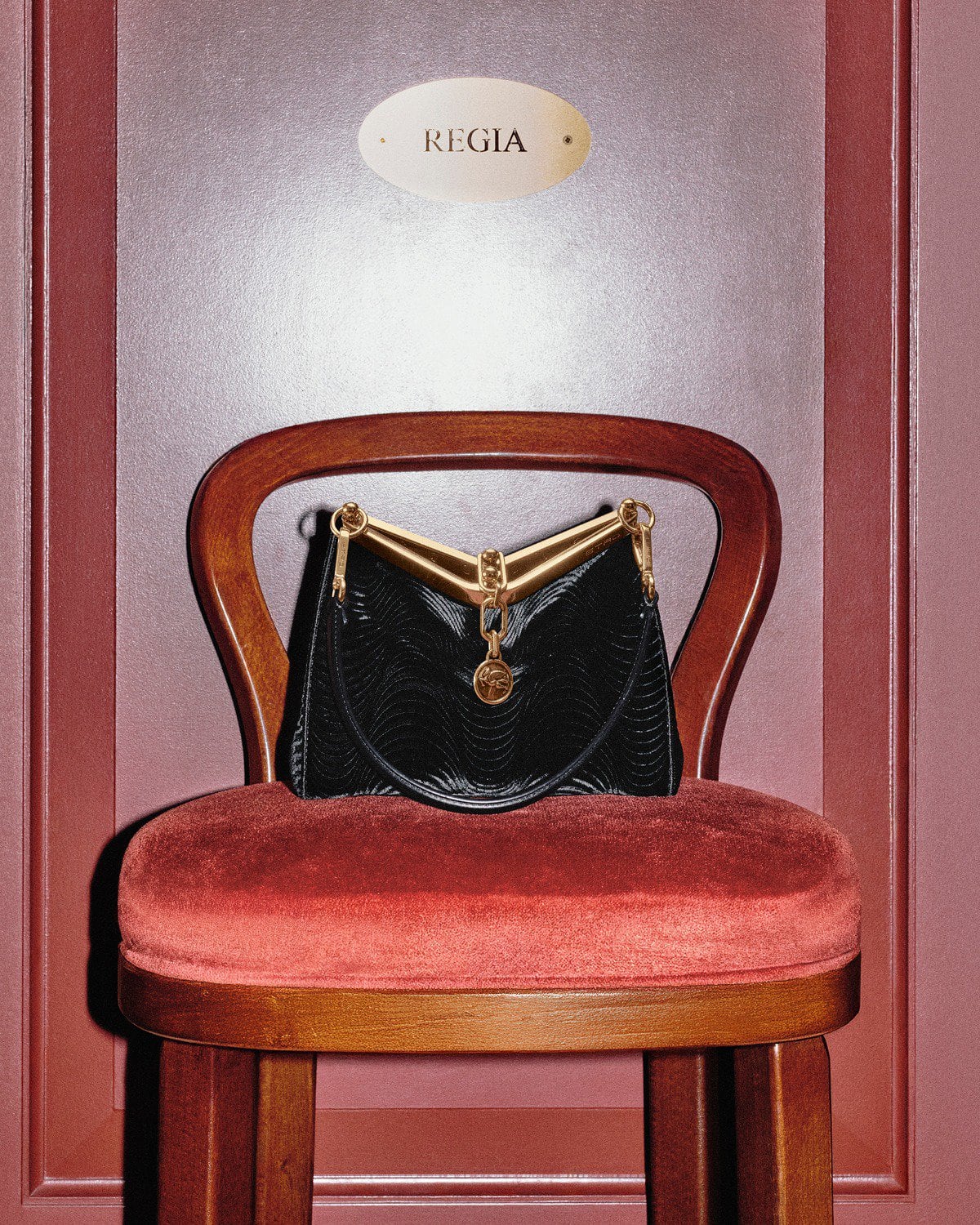 Black leather handbag with gold hardware resting on a red velvet chair against a warm red interior wall with an oval sign.