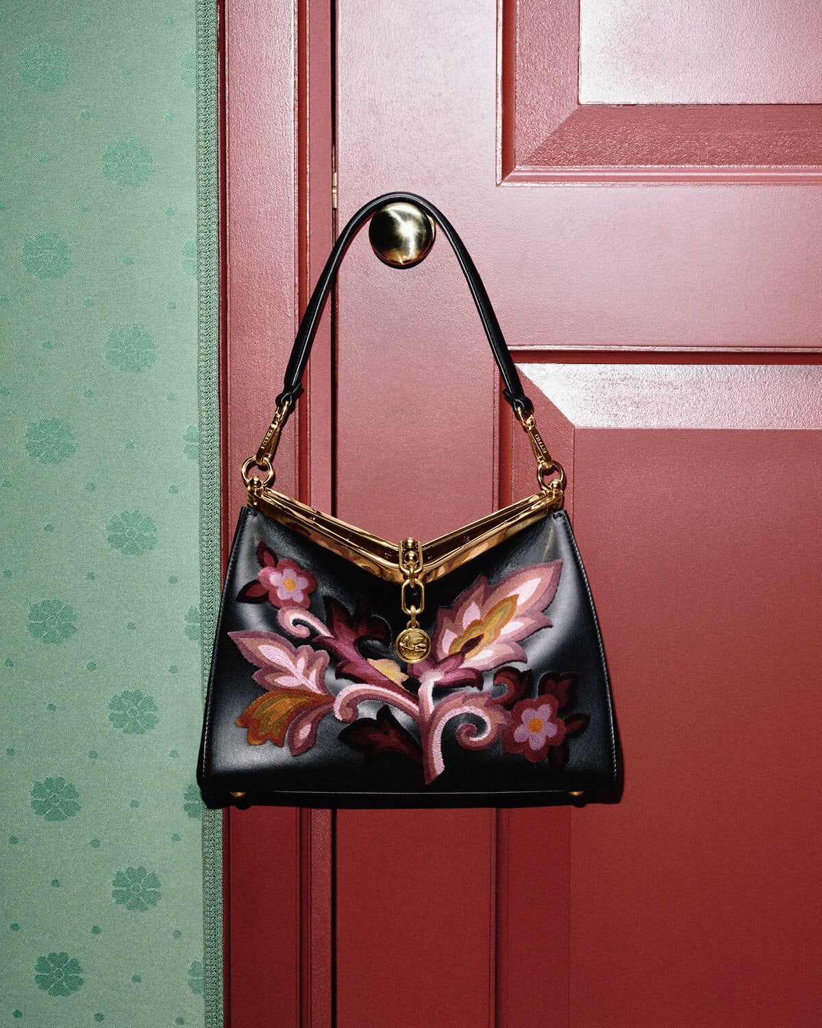 Black leather handbag with floral embroidery hanging from a brass doorknob on a red door against patterned green wallpaper.