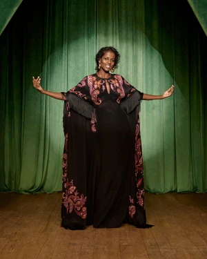 Model wearing a black patterned gown with fringe details standing on a stage with green velvet curtains.