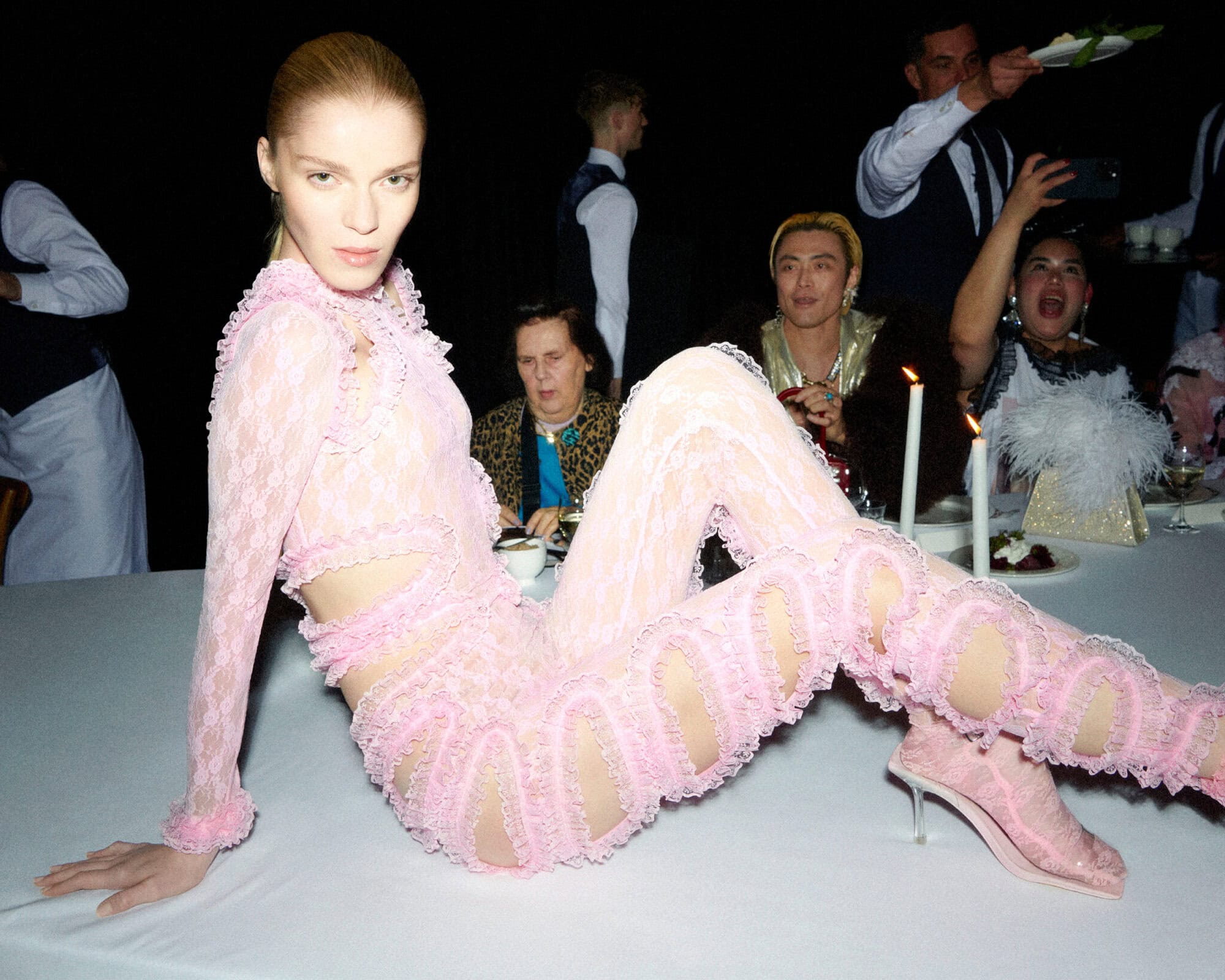 Model in a pink lace jumpsuit posing on a dining table with lit candles and servers in the background.