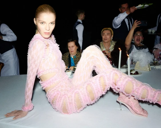 Model in a pink lace jumpsuit posing on a dining table with lit candles and servers in the background.