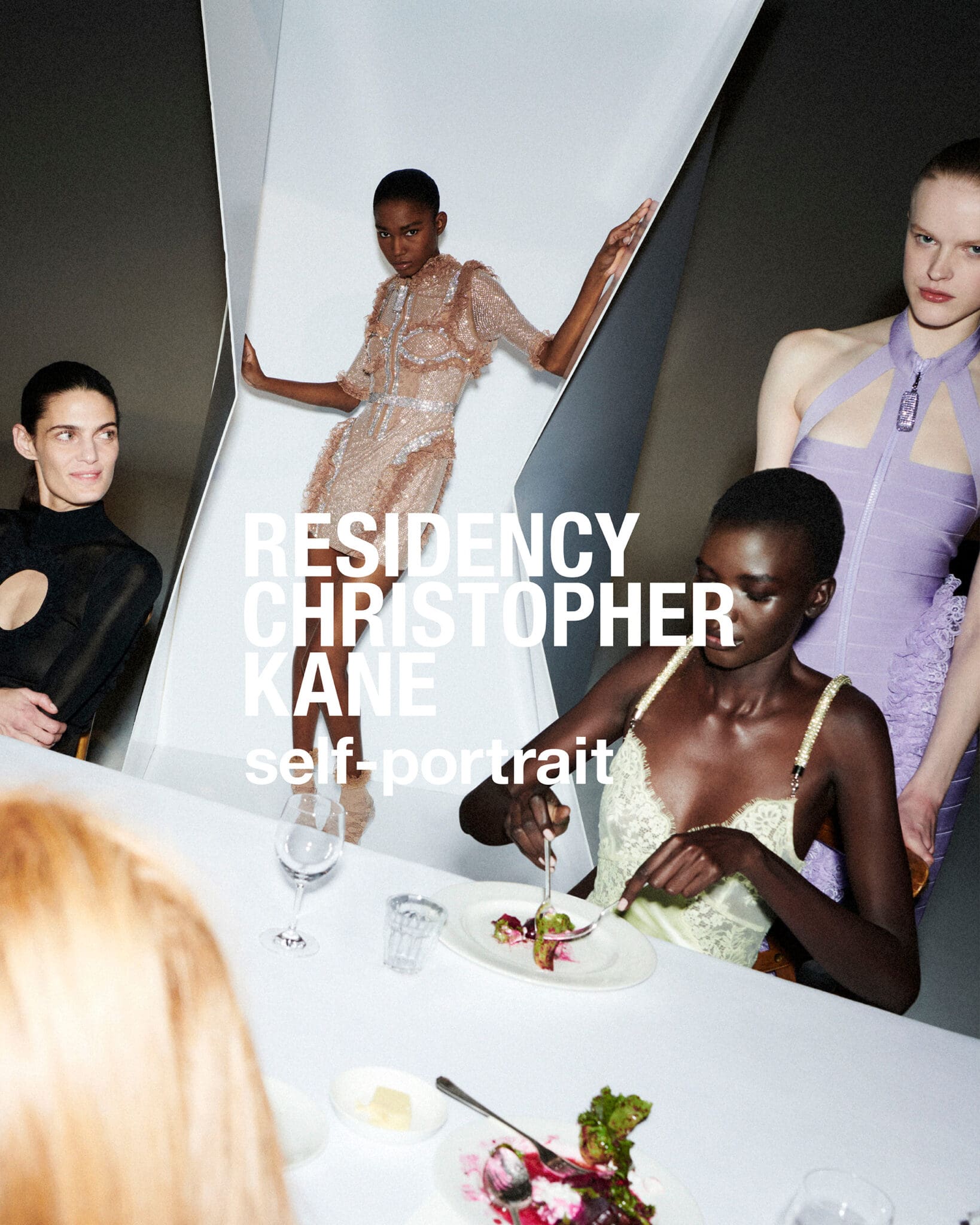Group of models gathered around a white dining table in a studio with graphic angular white geometric panels.