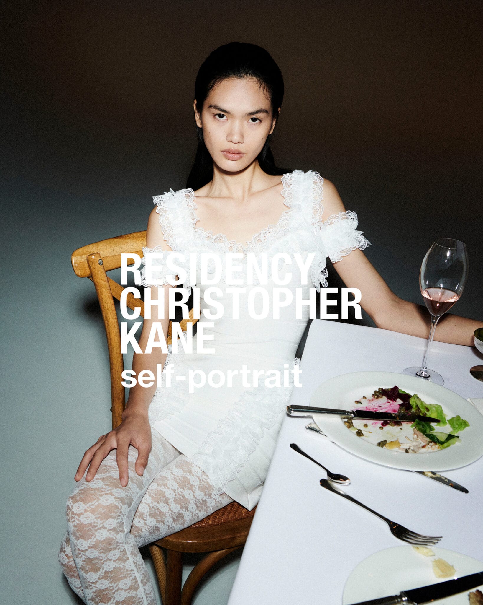 Model in a white lace dress seated at a dining table with plates and a wine glass against a dark background