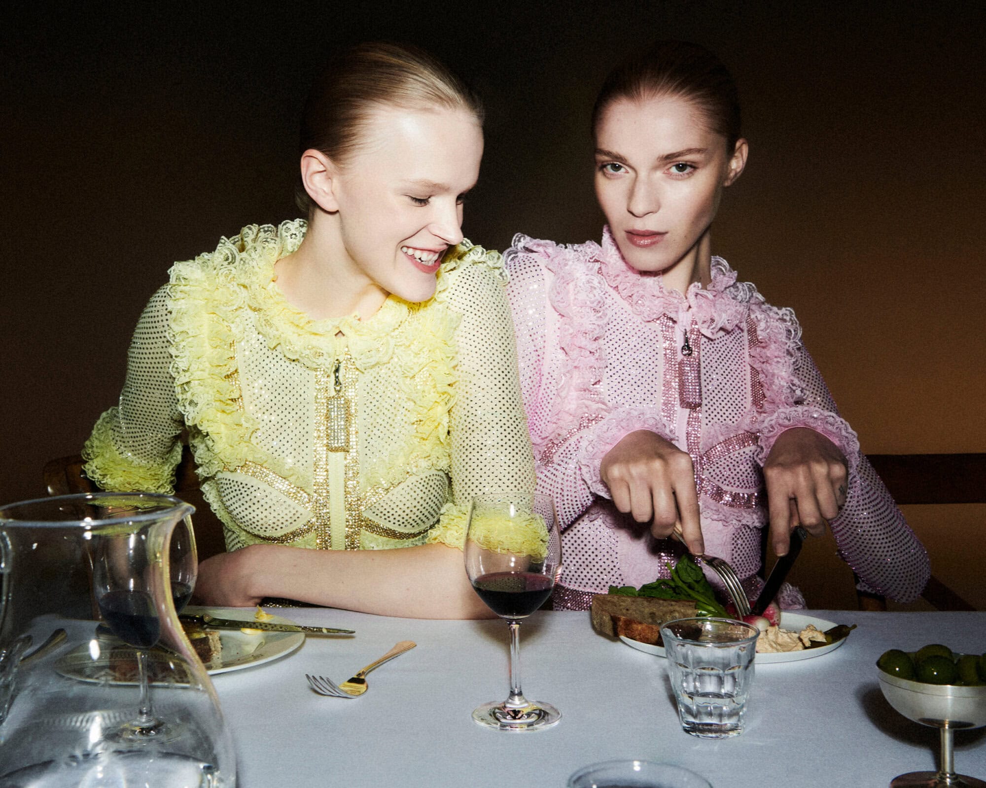Two women seated at a dining table, one wearing a yellow lace top and the other a pink lace dress, dining together.