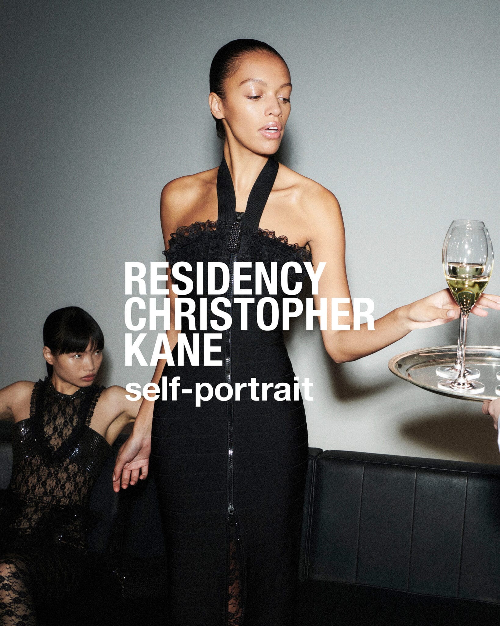 Model standing in a black halter dress holding a silver tray with wine glass against a grey wall
