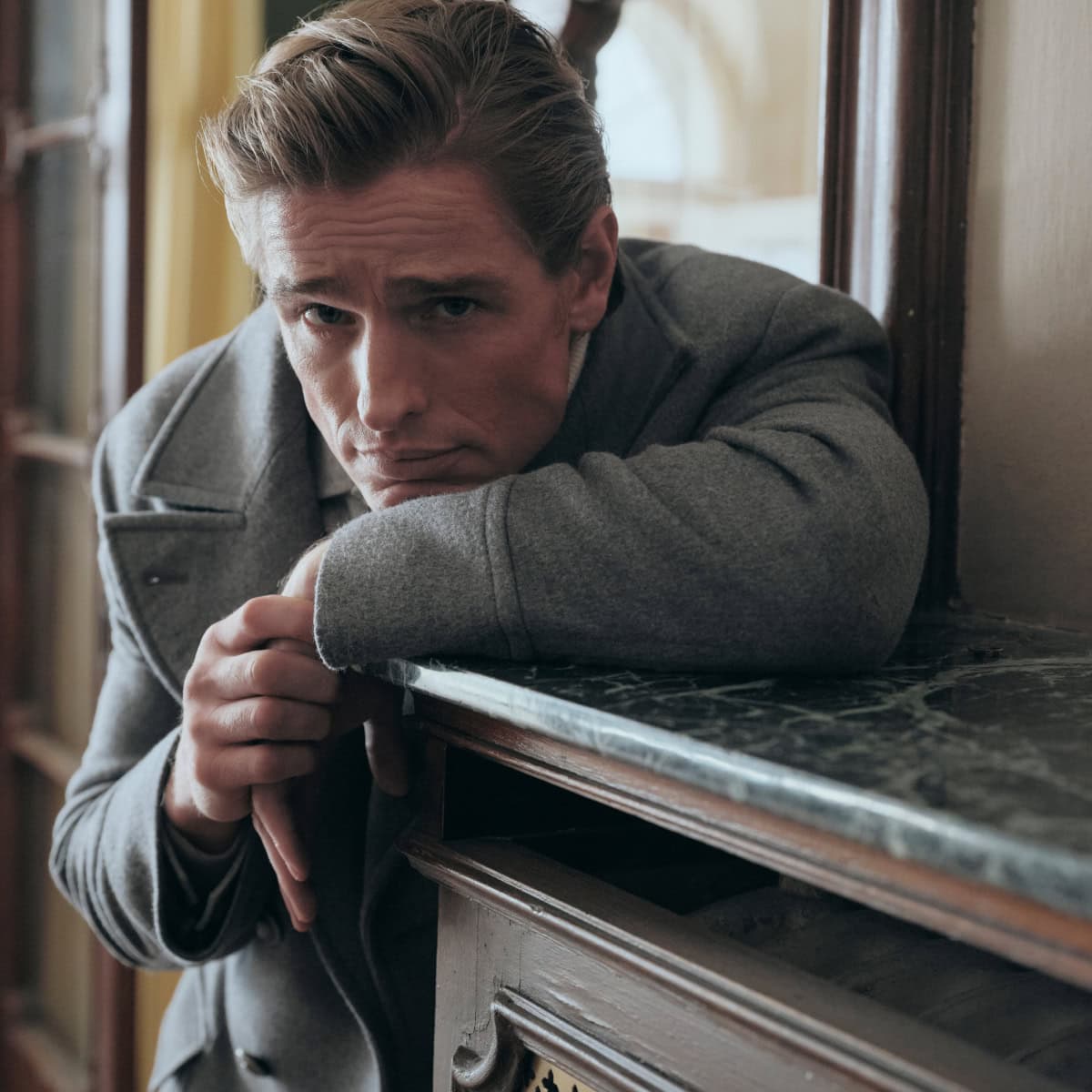 Man wearing a grey coat leaning on a marble-topped wooden console table in an interior space