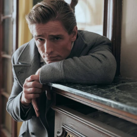 Man wearing a grey coat leaning on a marble-topped wooden console table in an interior space