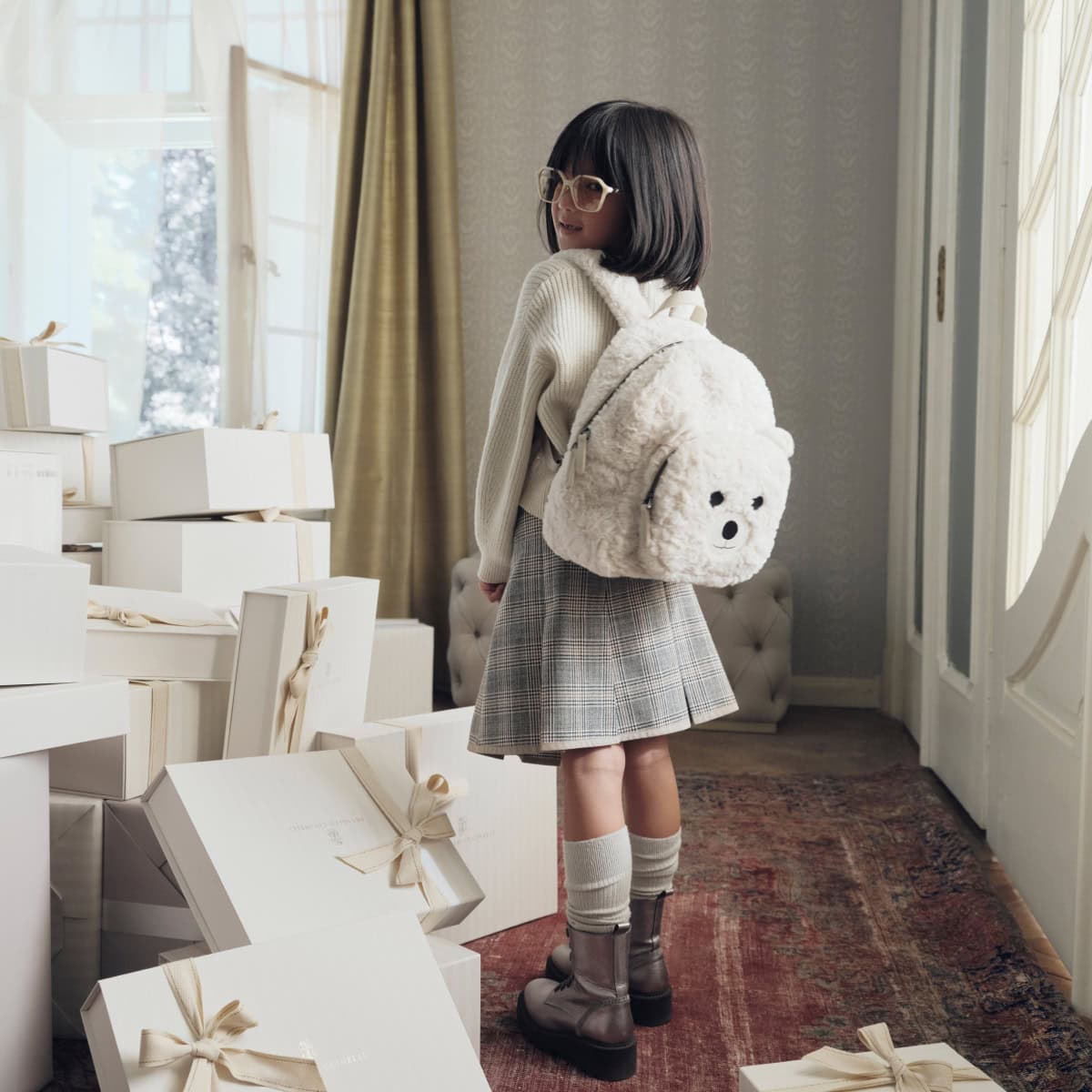 Young child wearing a cream sweater and plaid skirt with a fluffy white teddy bear backpack in a room with gift boxes