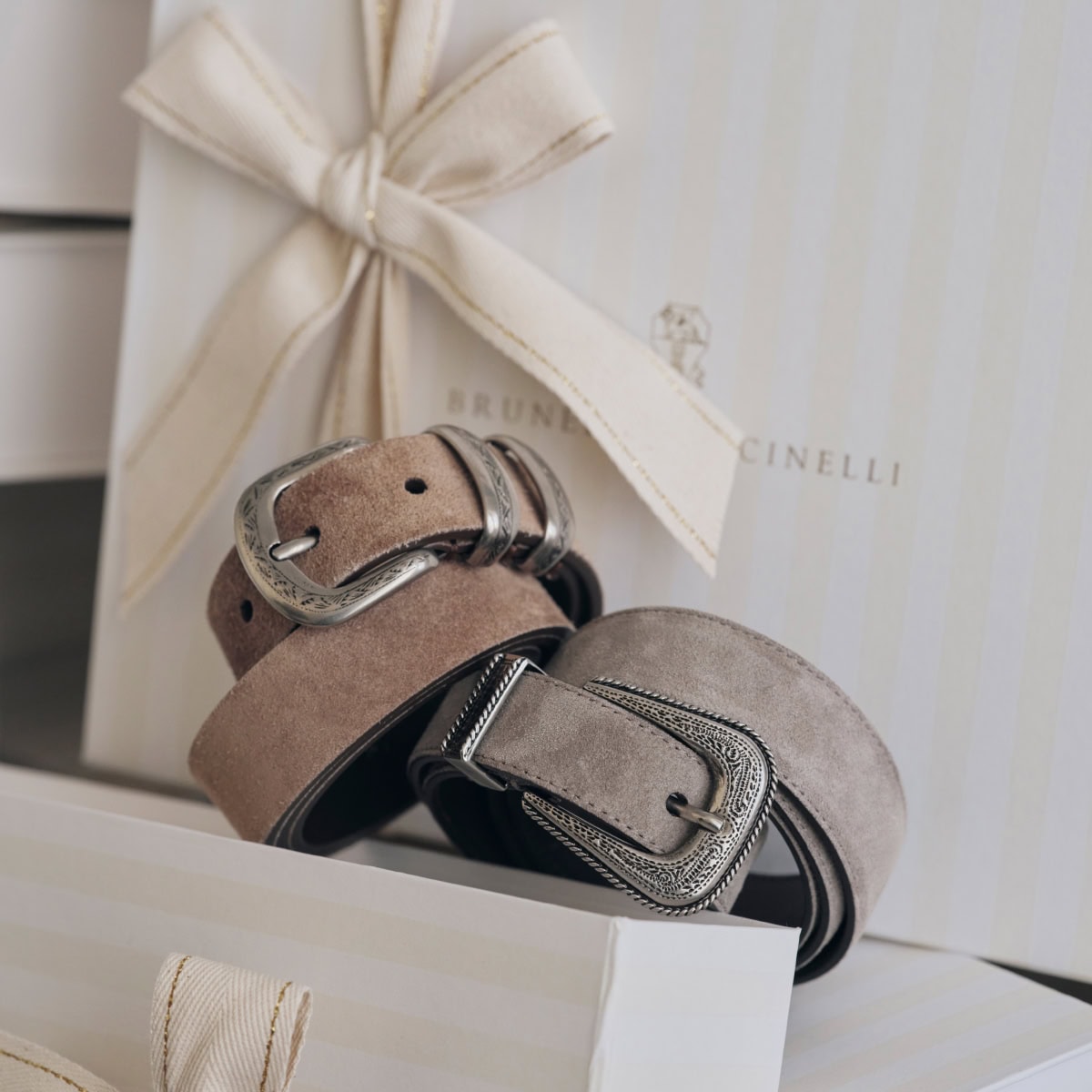 Two suede belts with ornate silver buckles resting against a gift box tied with a cream ribbon for Brunello Cucinelli