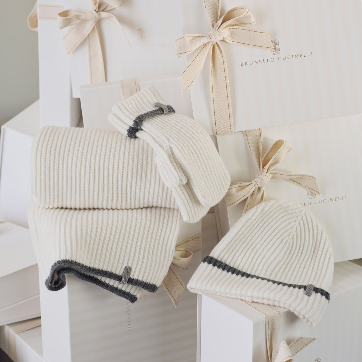 Stacked white knitwear set including scarf, beanie and mittens against stacked branded gift boxes with ribbon