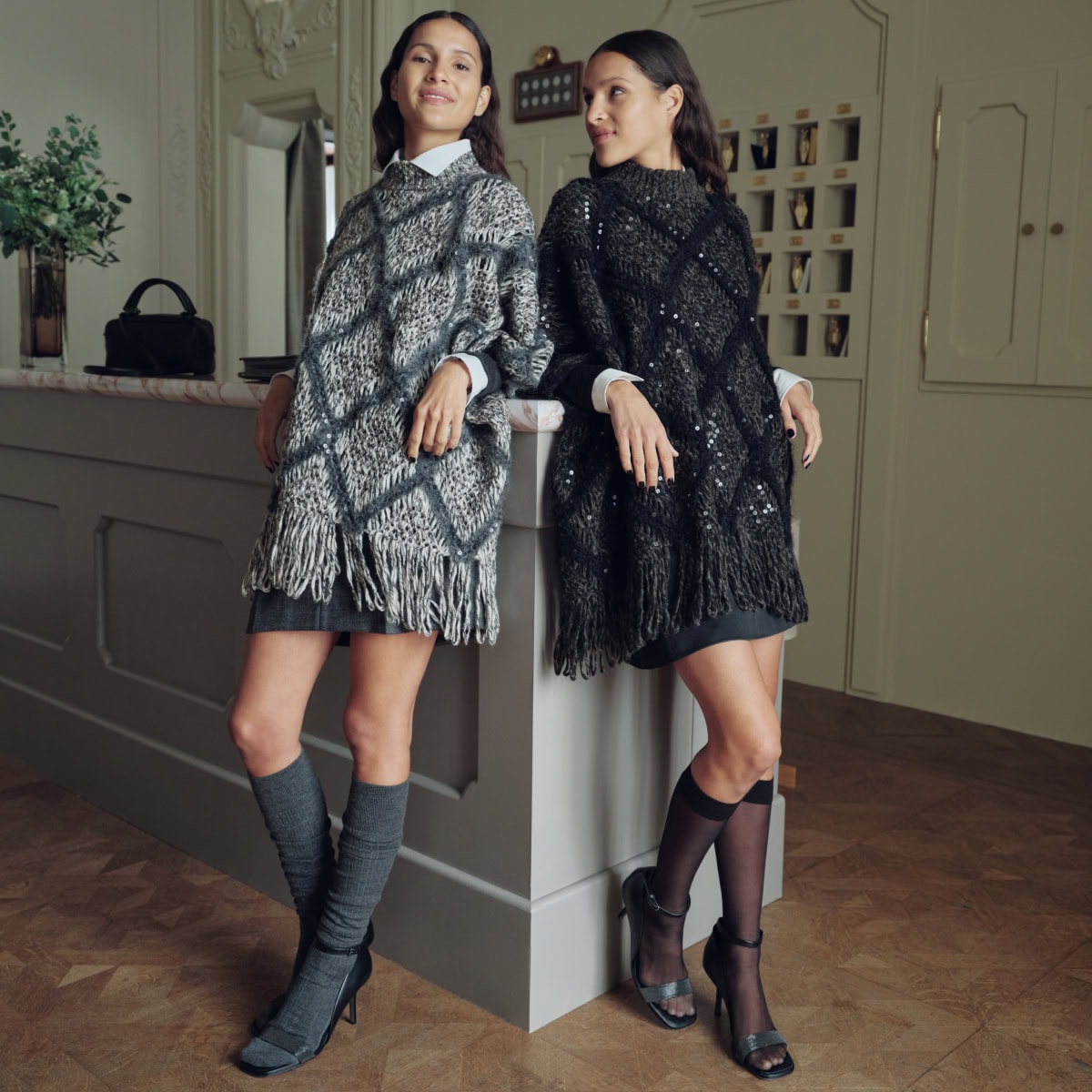 Two female models leaning against a counter wearing patterned knit ponchos and knee-high socks for Brunello Cucinelli