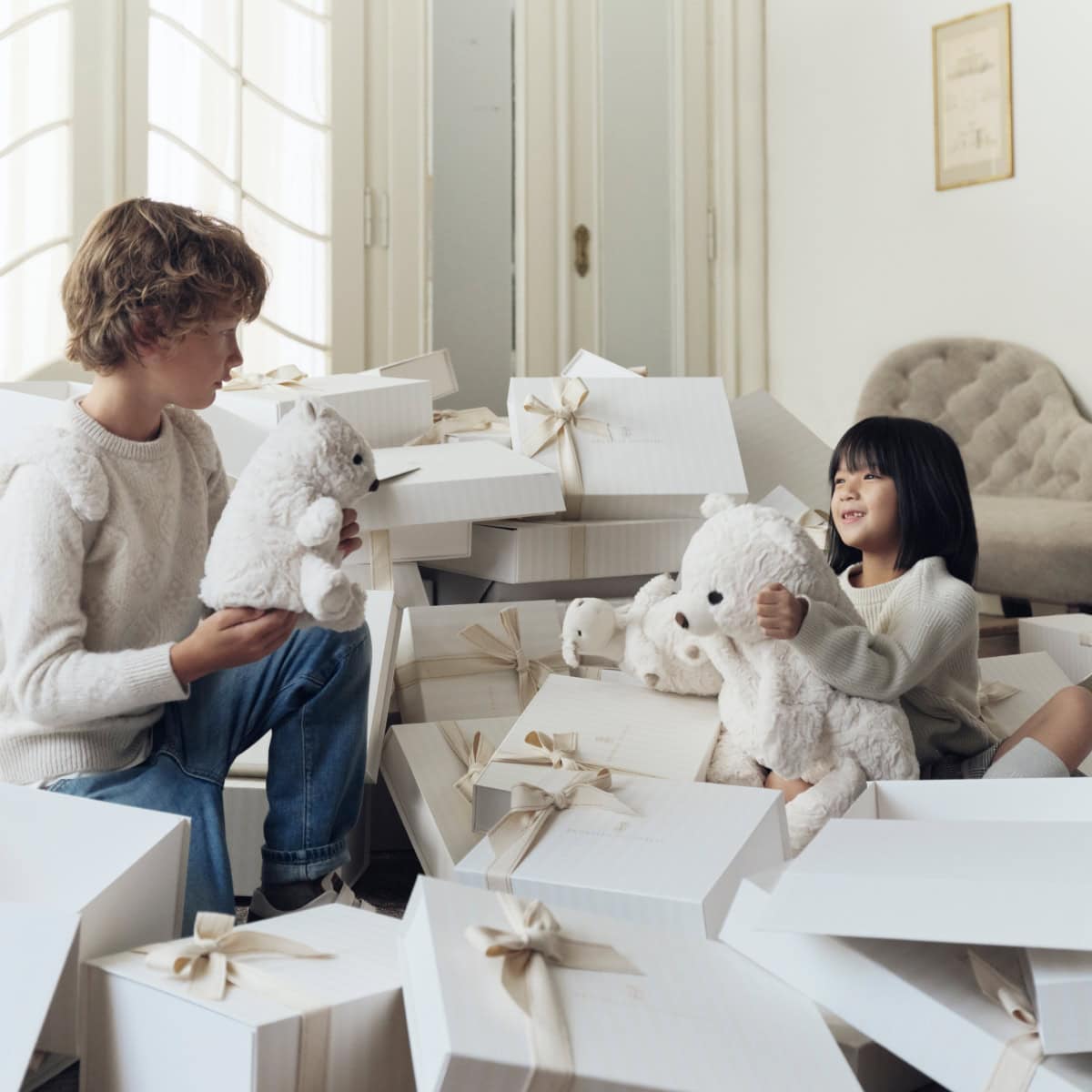 Two children sitting amidst stacked white gift boxes tied with ribbons, holding white plush teddy bears.