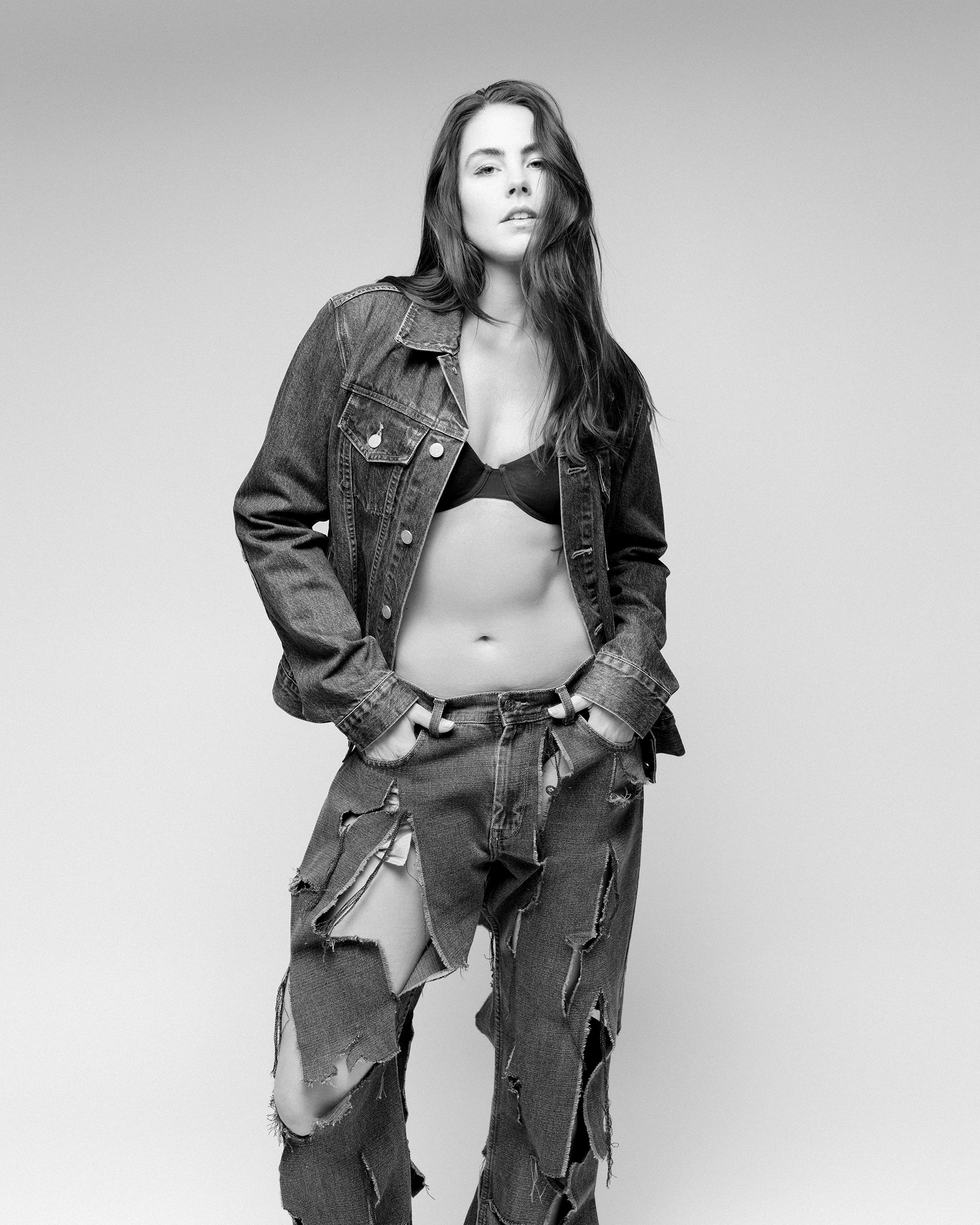 Black and white studio shot of an adult female model in torn denim jeans and jacket with a visible bra top and neutral facial expression.