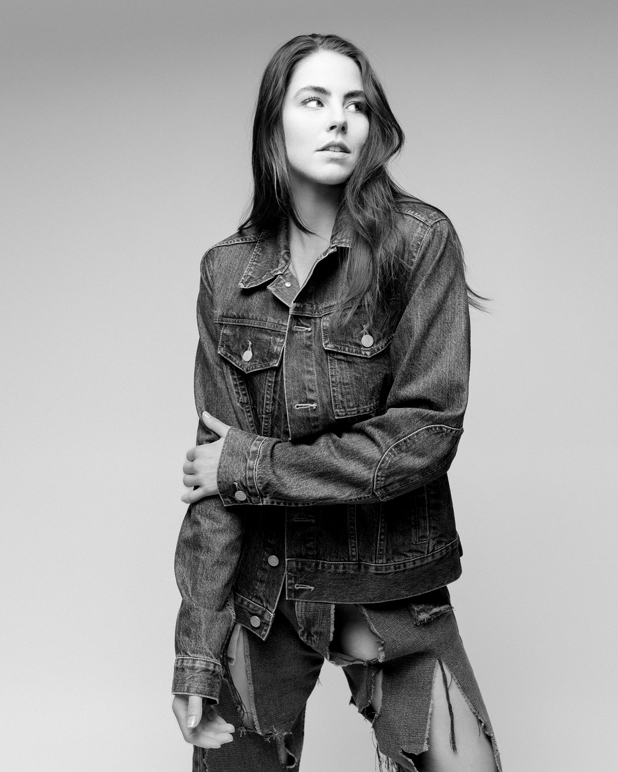 Black and white portrait of a young adult female in a denim jacket and ripped jeans looking left in a photo studio setting.