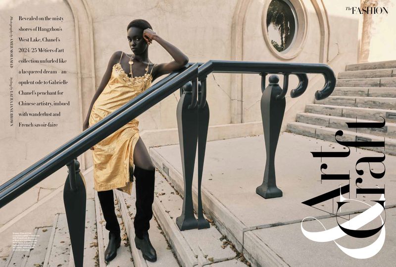 Fashion editorial shot of a Black model in a gold dress, leaning on a railing on stone steps with architectural details, bathed in soft light.