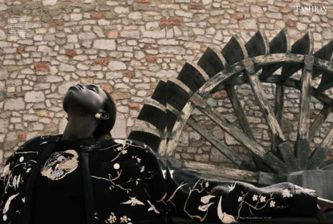 Fashion editorial portrait of an African woman in a patterned jacket looking up, set against a stone wall and a wooden water wheel.