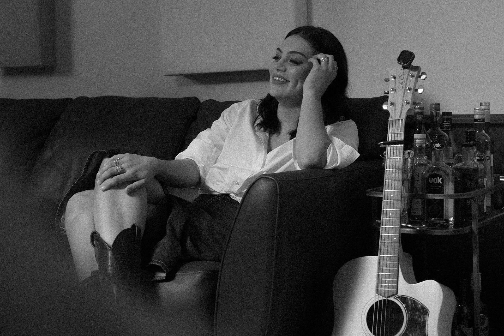 Thelma Plum seated on a leather sofa wearing a white button-down shirt and cowboy boots with an acoustic guitar and bar cart nearby.