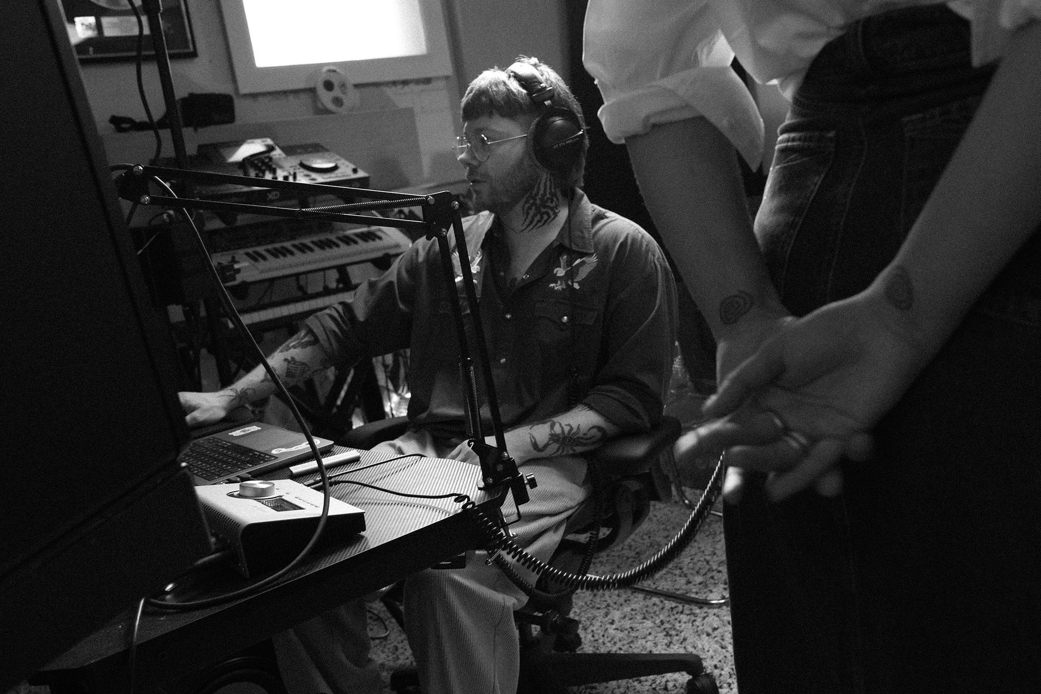 Man wearing headphones seated at a mixing desk in a recording studio with another person standing in the foreground