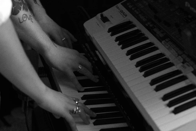 Close-up black and white image of hands playing electronic keyboard synthesizer keys with rings and tattoos visible.