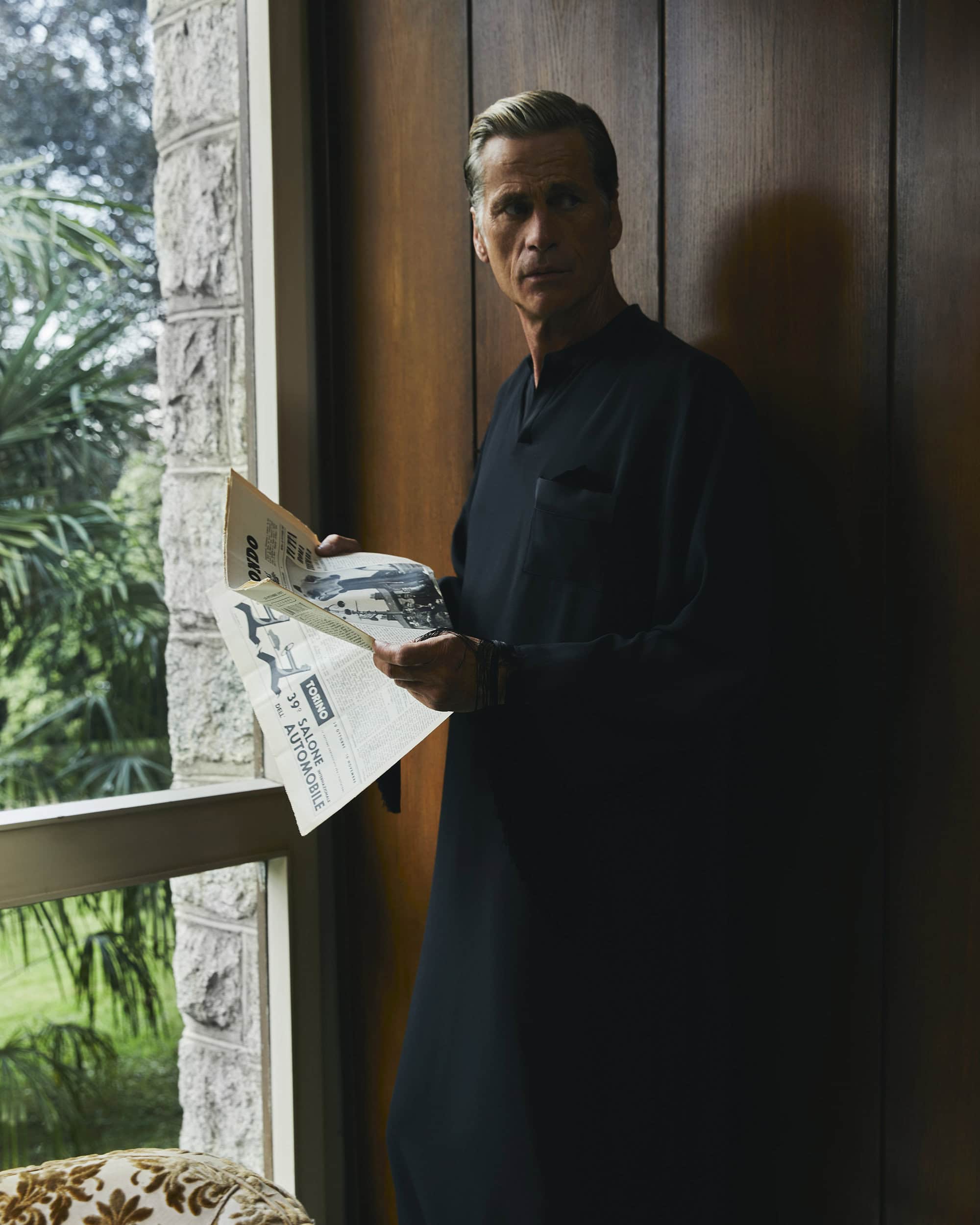 Middle-aged man in a black tunic holding open a newspaper near a window with foliage outside for Taller Marmo ad campaign.