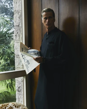 Middle-aged man in a black tunic holding open a newspaper near a window with foliage outside for Taller Marmo ad campaign.