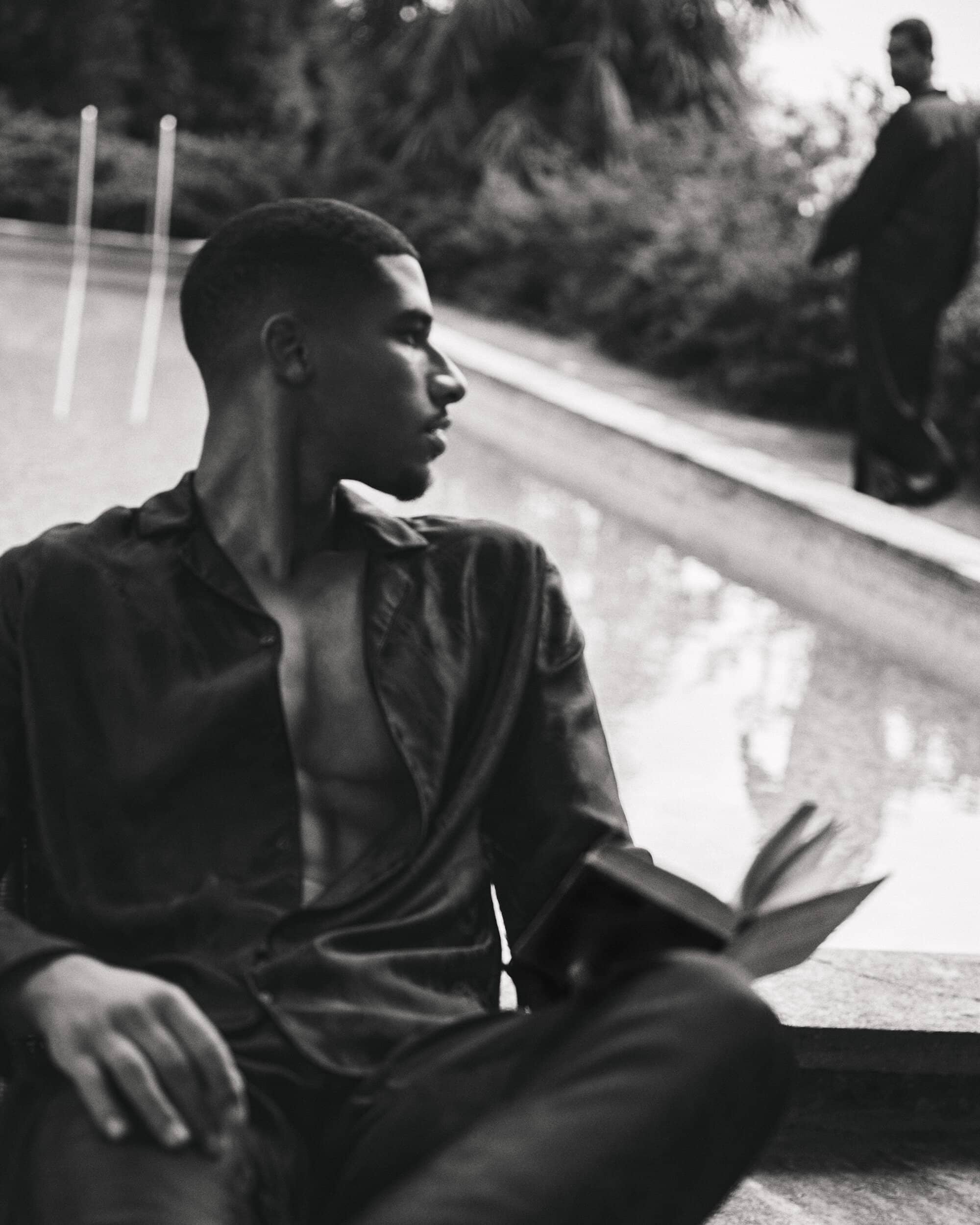 Black and white portrait of a man holding an open book near a pool with another figure in the background for Taller Marmo
