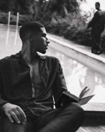 Black and white portrait of a man holding an open book near a pool with another figure in the background for Taller Marmo