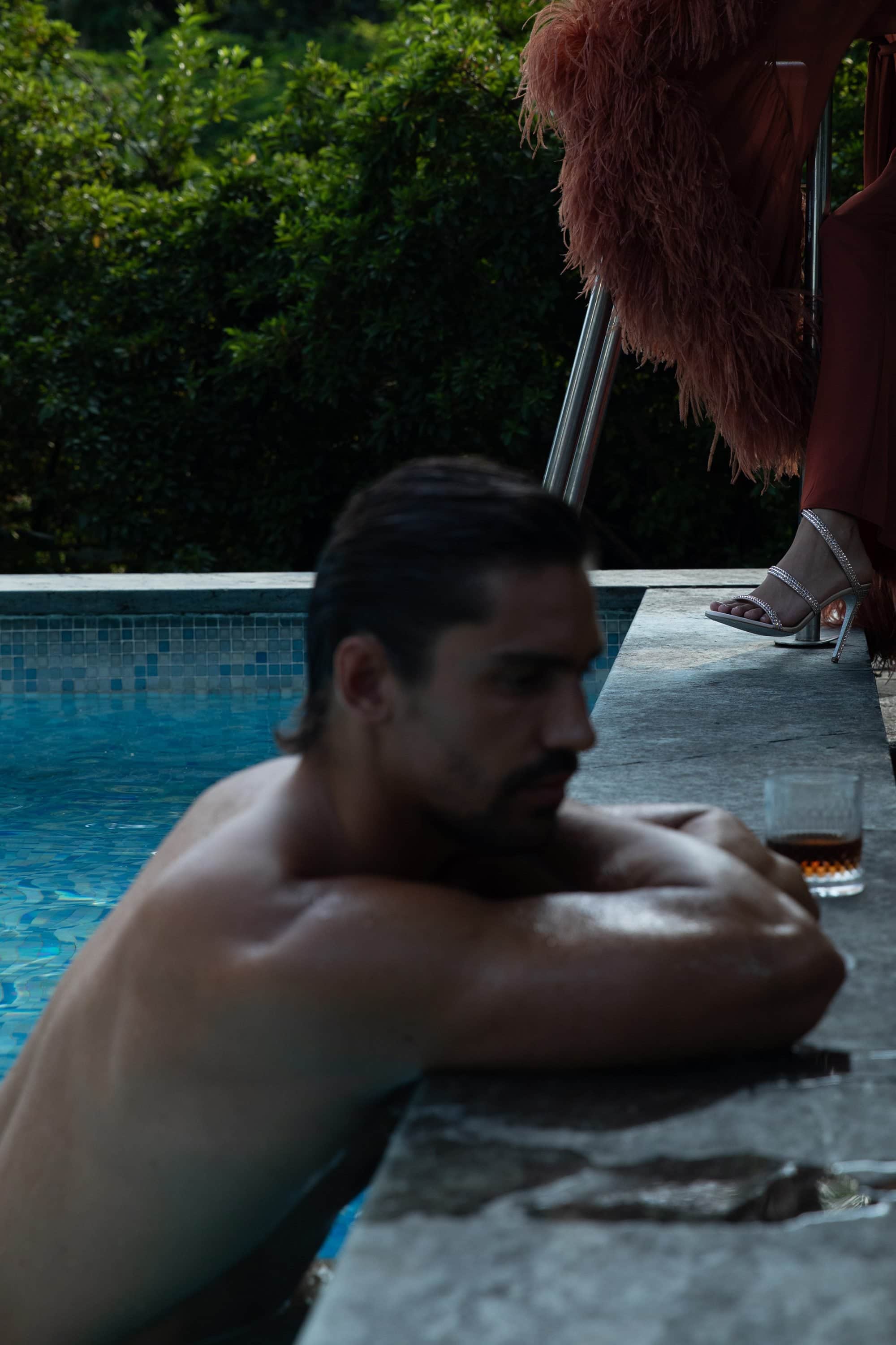 Shirtless man resting arms on edge of outdoor swimming pool with a glass of whiskey nearby