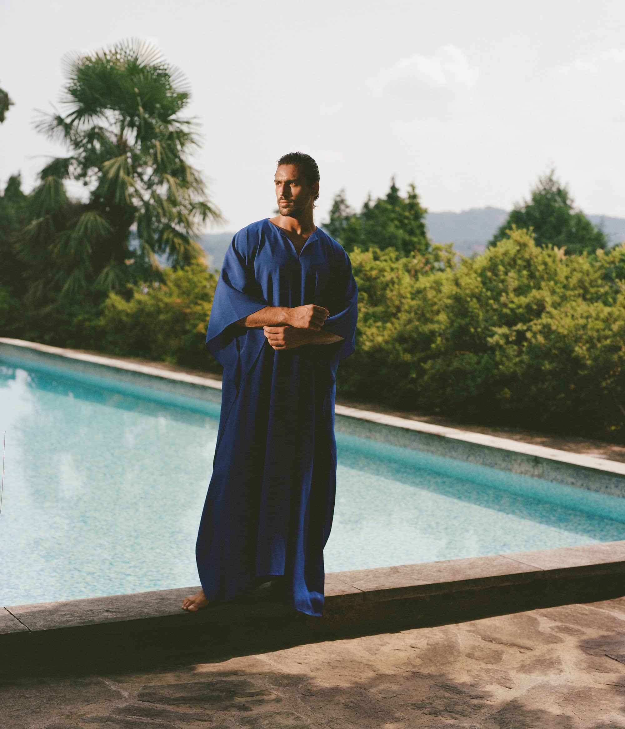 Man wearing a long blue kaftan standing by an outdoor swimming pool with palm trees in the background for Taller Marmo