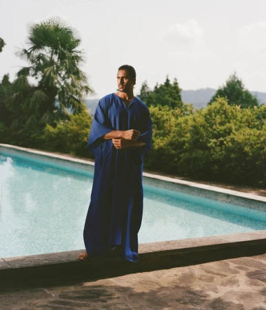 Man wearing a long blue kaftan standing by an outdoor swimming pool with palm trees in the background for Taller Marmo