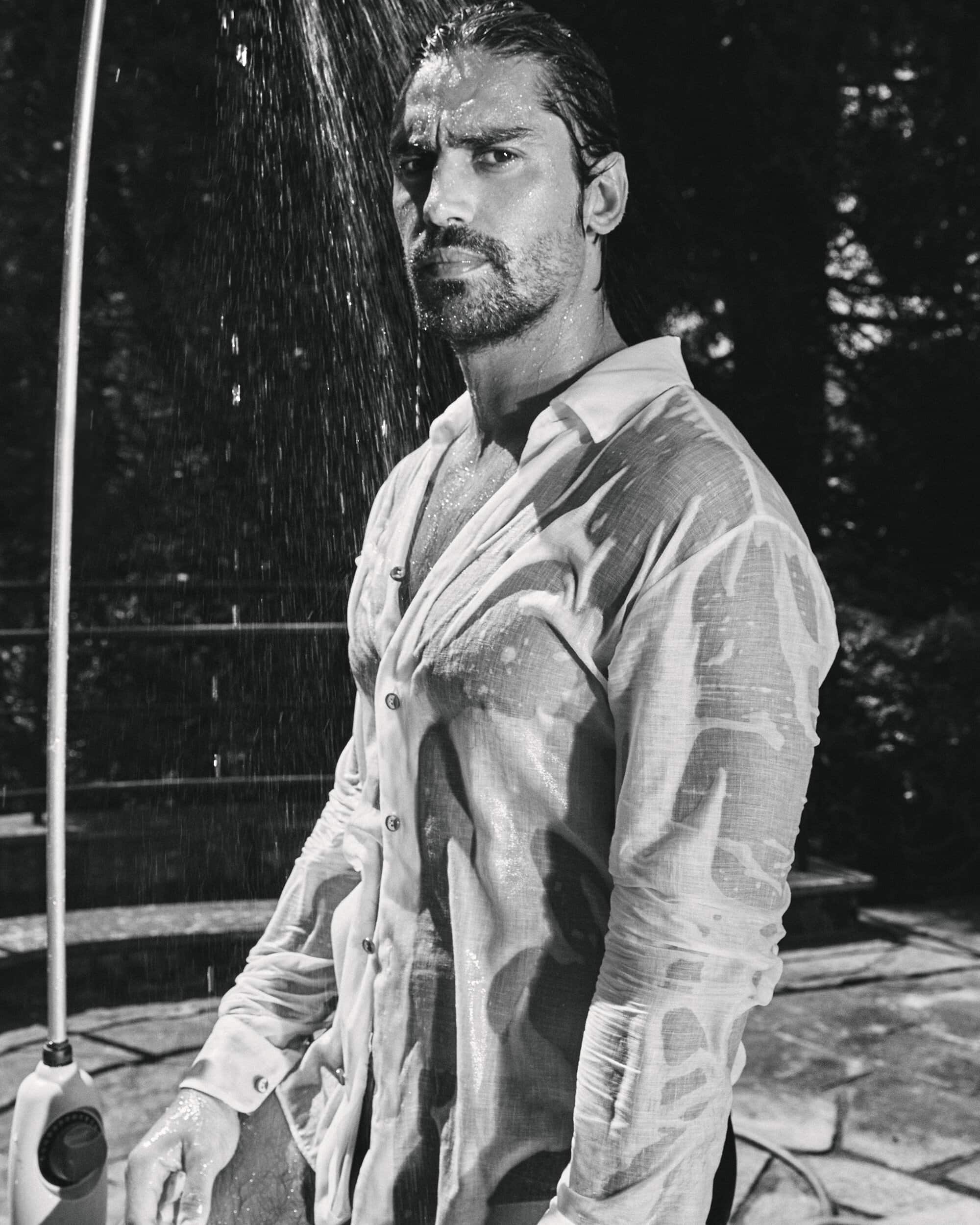 Monochrome portrait of a man with long hair in a wet white buttoned shirt standing outdoors beneath a shower stream.