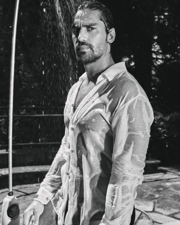 Monochrome portrait of a man with long hair in a wet white buttoned shirt standing outdoors beneath a shower stream.