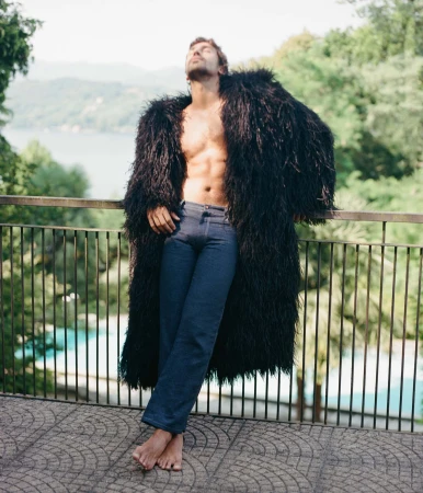 Man wearing a long black fluffy coat and denim jeans leaning against a balcony railing overlooking a scenic lake landscape.