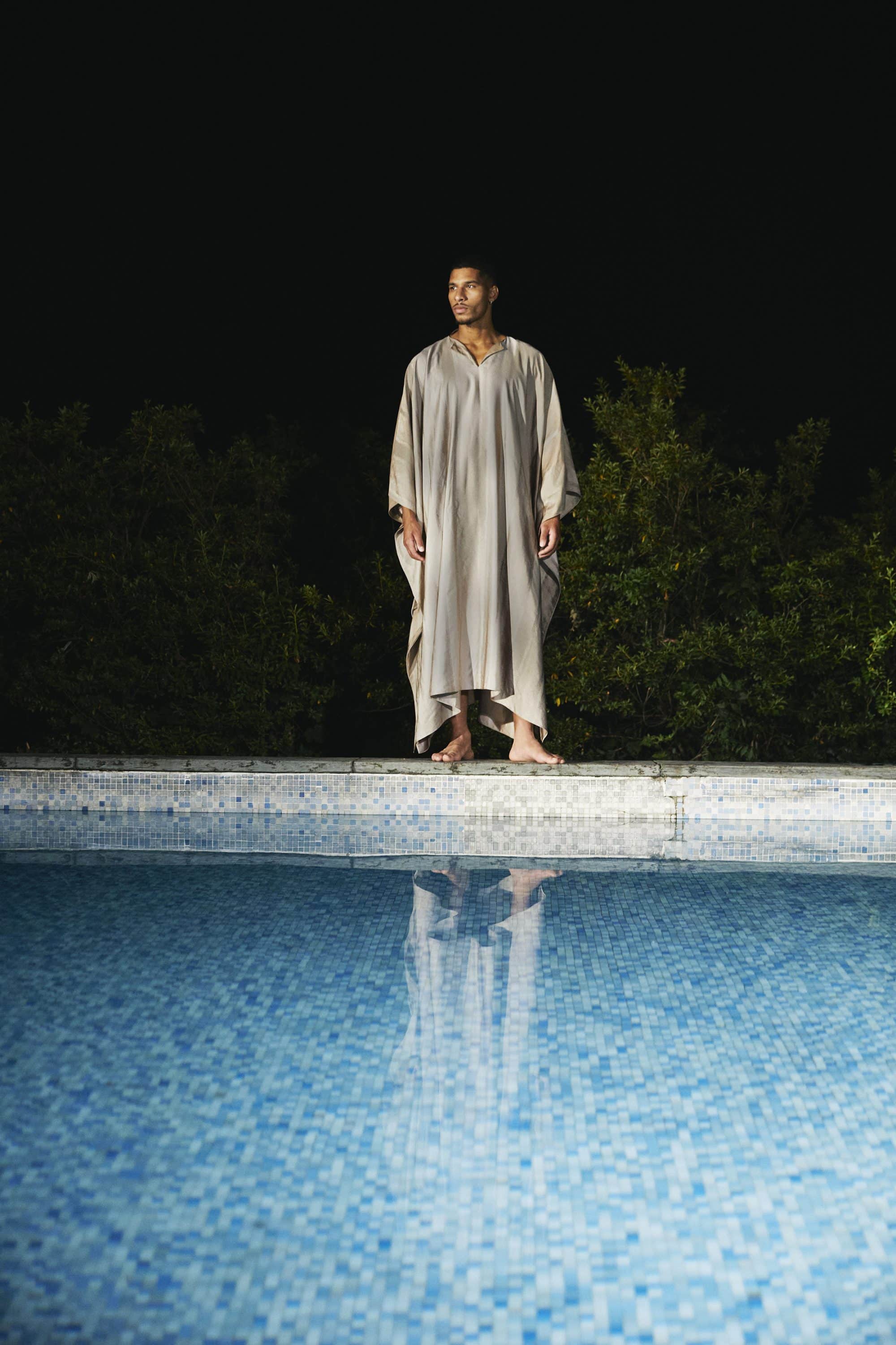 A man wearing a long cream kaftan standing barefoot by a swimming pool at night, with dark foliage behind him.