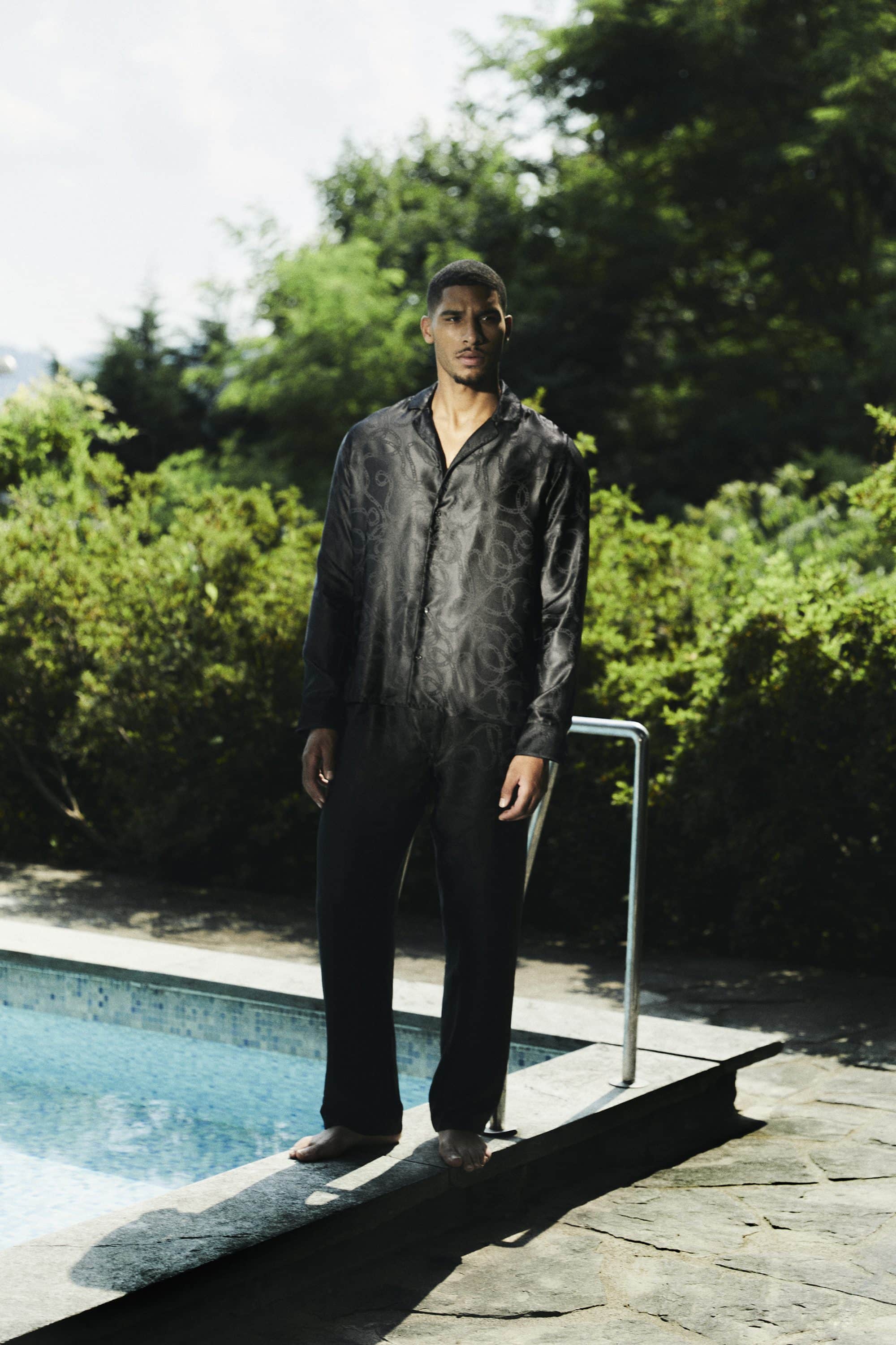 Man standing by an outdoor swimming pool wearing a black patterned silk shirt and matching trousers.