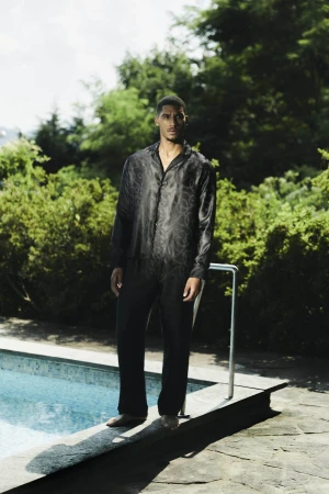 Man standing by an outdoor swimming pool wearing a black patterned silk shirt and matching trousers.