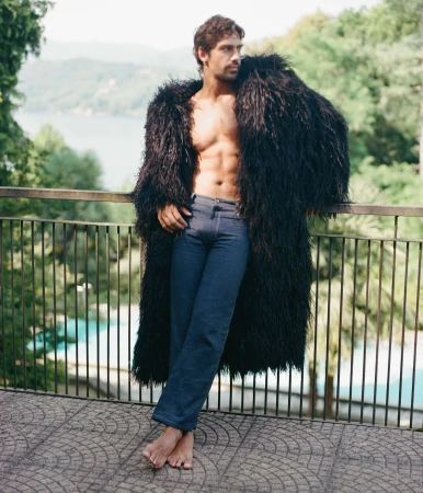 Man wearing a long black fluffy coat and denim jeans leaning against a balcony railing outdoors