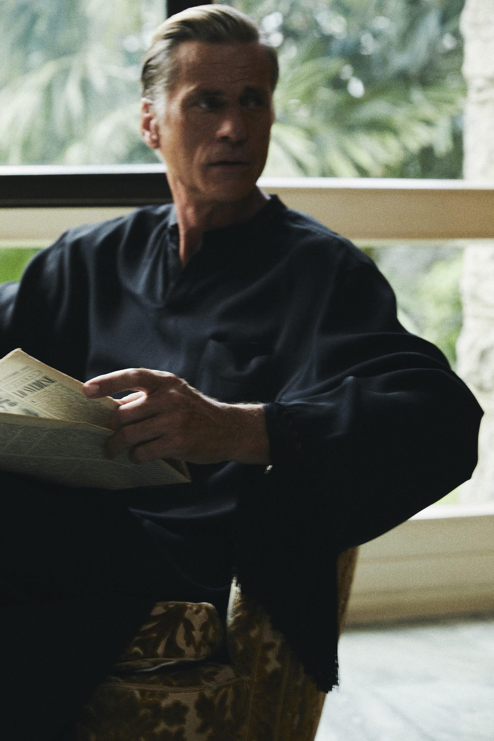 Middle-aged man wearing a black long-sleeve shirt seated in an armchair holding a newspaper