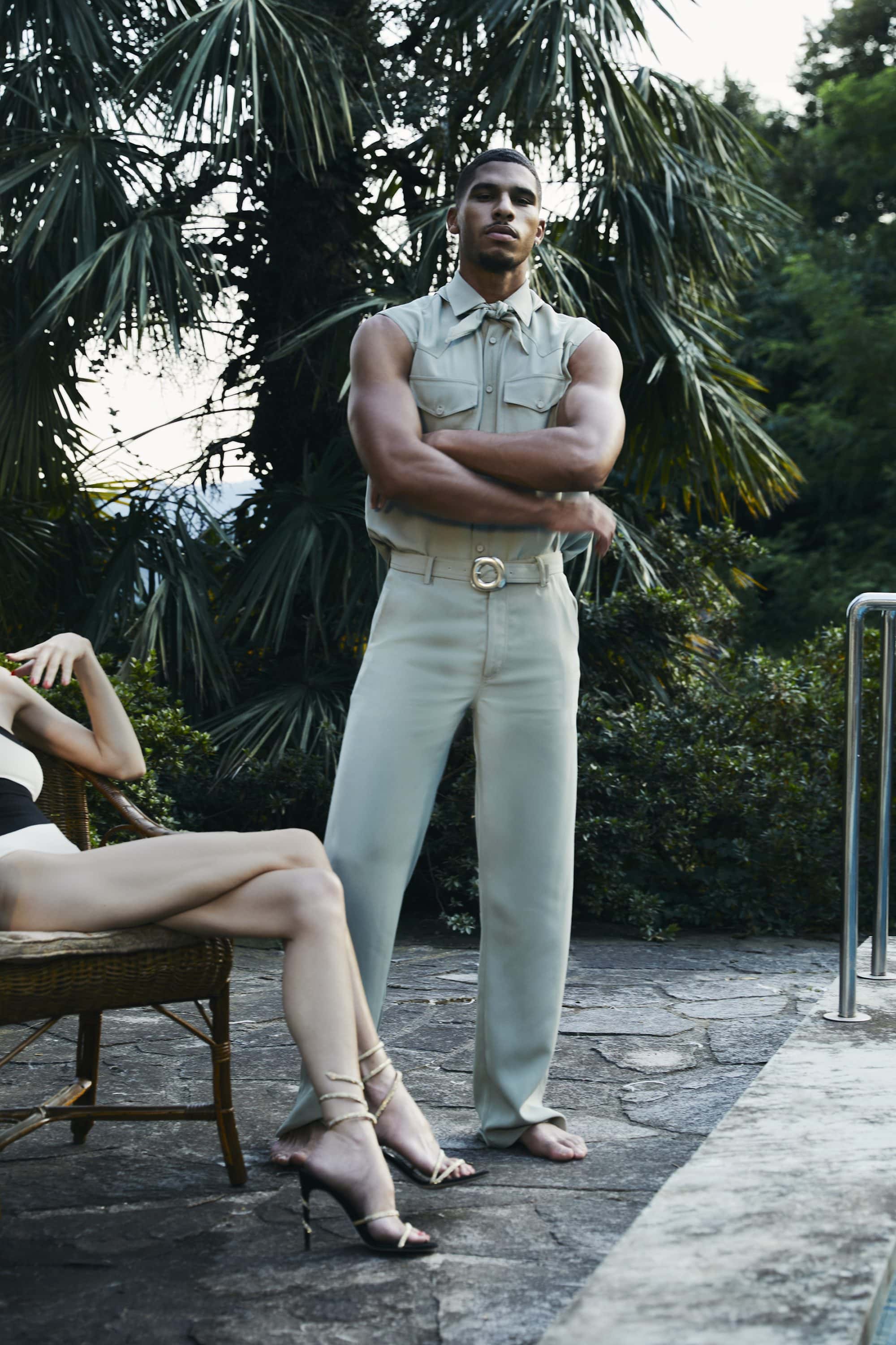 Man standing barefoot with arms crossed in pale green clothing outdoors near a pool with palm foliage in background